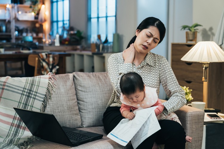 Ilustrasi Bunda Pekerja Alami Burnout portrait asian working mother holding baby is having tension in her neck. millennial mom spraining her neck while looking after her child. lovely toddler is looking at document in her mother’s hand