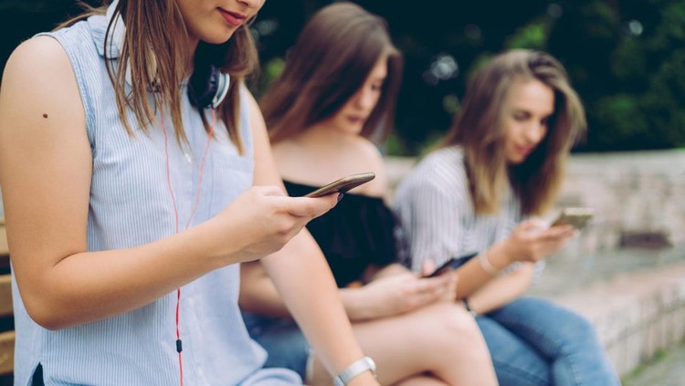 Three beautiful teenage girlfriends looking at the phone, sitting on the bench, downtown.