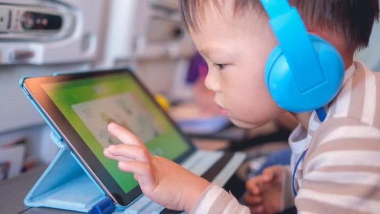 Cute Asian 2 - 3 years old toddler boy child wearing headphones using tablet pc watching cartoons / playing game during flight on airplane. Happy Flying with children concept, soft & selective focus