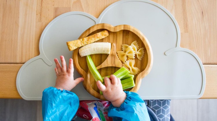Baby hands grabbing food from plate (bamboo) with finger food (cucumber, pasta, banana) for babies in top view, showing the concept of baby-led weaning, nutrition without pureed food