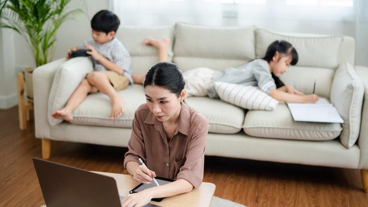 Asian mother working with laptop while her son and daughter are playing in the living room.