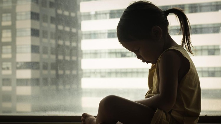 A litte girl sitting next to a window with her head down in sadness. Feeling depressed and hurt.
