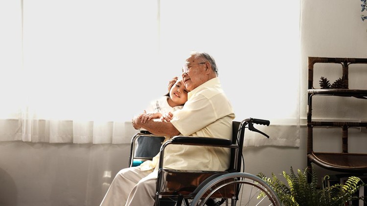 Asian senior old man hugging granddaughter come visitor at home, Grandfather sitting wheelchair on sofa in living room, Caregiver mature concept.