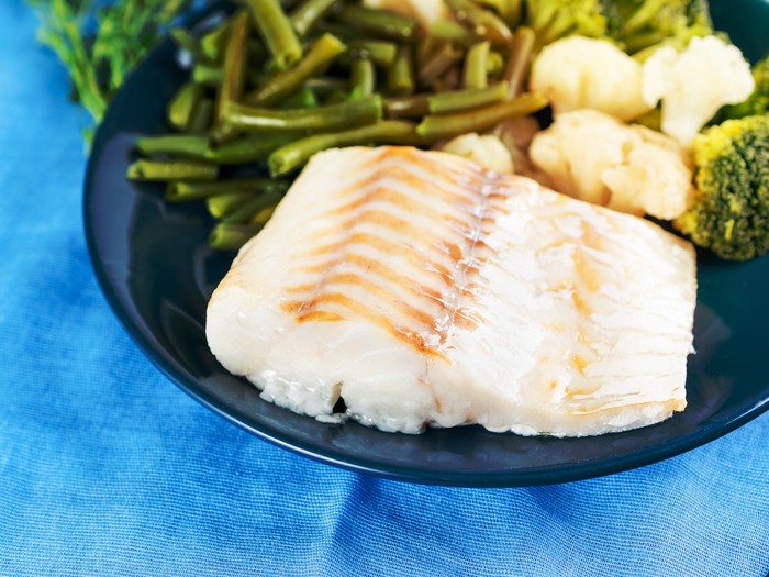Fillet of boiled sea fish cod with broccoli, green beans and cauliflower on a blue plate, side view, close. Healthy diet dish for proper nutrition
