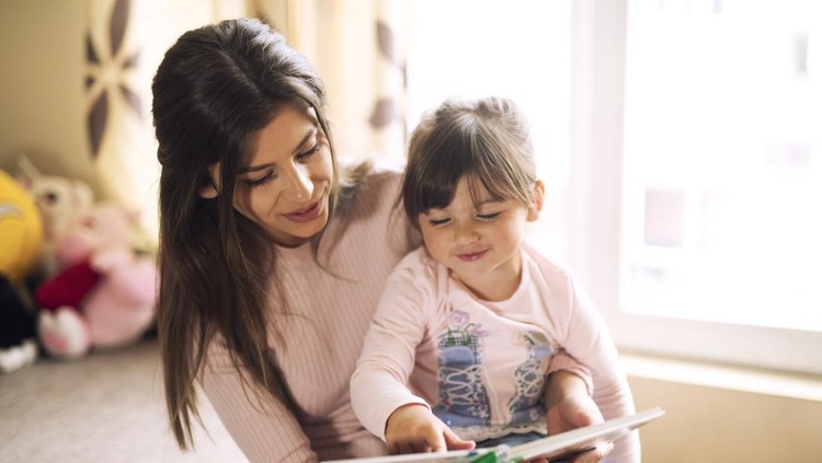 Happy mother and daughter cuddling, reading book in sunny living room
