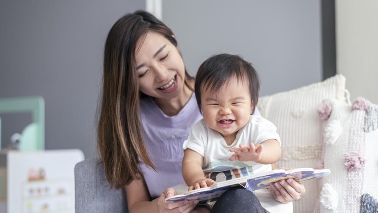 A beautiful young mother of Asian descent sits on the couch at home and affectionately holds her cute baby in her lap while reading a book to the child. The happy baby is looking with interest and delight at the book.