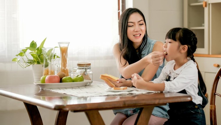 Mother take care and give bread to her daughter to eat in kitchen. Concept of good relationship in family and new normal lifestyle at home.