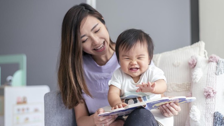 A beautiful young mother of Asian descent sits on the couch at home and affectionately holds her cute baby in her lap while reading a book to the child. The happy baby is looking with interest and delight at the book.