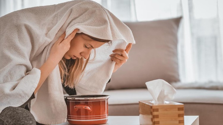 Health care, flu, hygiene and people concept - Photo of sick brown hair woman at home inhaling over a bowl. Rhinitis treatment at home by inhalation. Young woman inhaling chamomile tea steam as an alternative therapy or traditional cure. Young woman inhales steam to treat a blocked nose during the day.