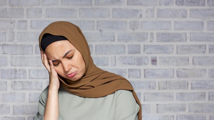 a young asian woman in muslim attire, feeling upset disapointment, worry, sick, headache with a concrete wall as background