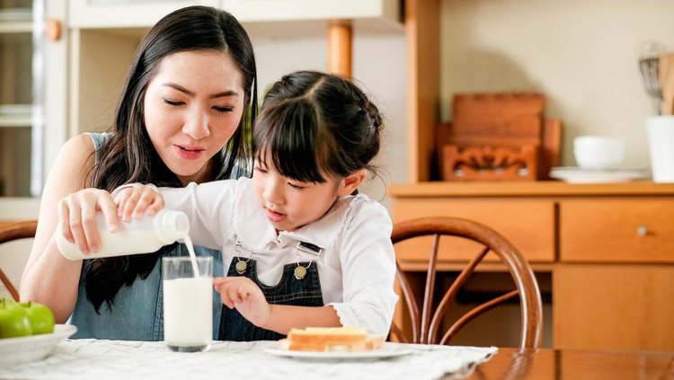 Asian mother take care her daughter pour milk to a glass on the table in house kitchen during breakfast meal. Main focus is the mother.