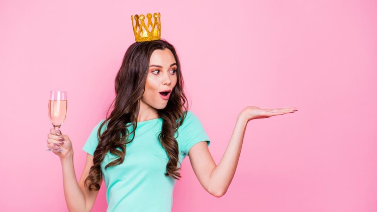 Sweet sixteen! Curly brunette girl holds a glass of beverage in one hand and enthusiastically looks at an invisible object with open mouth isolated on shine pink background with copy space for text