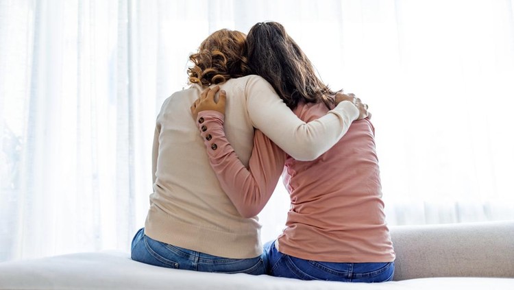 Rear back view of a mother and daughter embrace sitting on bed at home, older sister consoling younger teen, girl suffers from unrequited love share secrets trustworthy person relative people concept