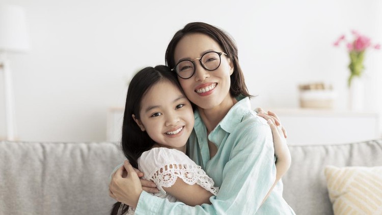 Happy little Asian girl and her loving grandmother hugging and looking at camera on couch at home. Portrait of granny and grandchild embracing, expressing love and affection indoors
