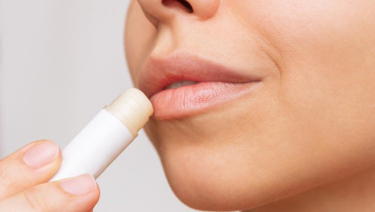 Cropped shot of a young caucasian beautiful woman applying a hygienic lipstick on her lips on a gray background. Moisturizing chapstick for dry lips