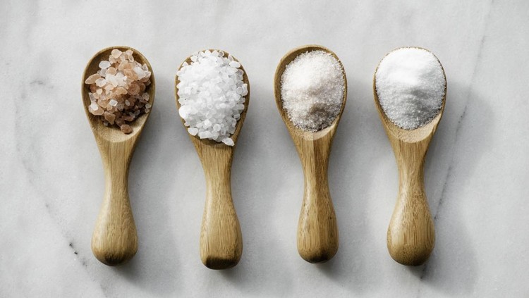 Various salt types in 4 wooden spoons on white marble.