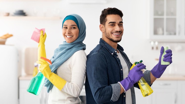 Cheerful middle-eastern handsome husband and pretty wife in hijab cleaning kitchen together, wearing rubber gloves, holding cleaning sprays, sponges and dust clothes, standing back to back and smiling