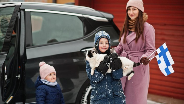 Kids with cat on hands against car in the yard of house at winter. Mother hold flag of Finland.