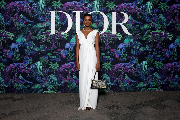 Simone Ashley dari serial Sex Education (2020) tampil elegan mengenakan gaun putih dan kalung. Statement datang dari tas berhiaskan corak bunga. Foto: Getty Images/Courtesy of Dior