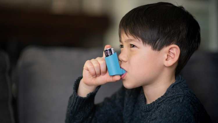 A little boy is sitting in a living room. He is using puffer because he suffers from asthma.