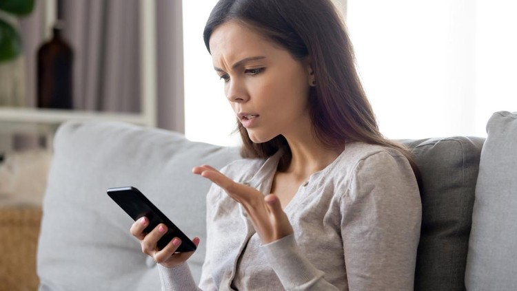 Upset young woman having problem with mobile phone, irritated girl reading bad news in message or email, looking at screen, discharged or broken mobile device, sitting on sofa at home