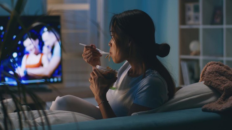 1342504764 Asian woman eating takeaway noodles and watching TV while sitting on couch and having dinner in evening at home