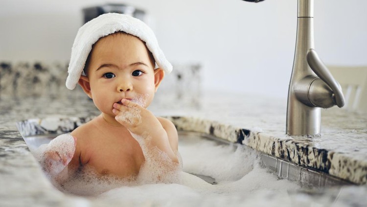 A 9 month old baby taking a bath in a kitchen sink.