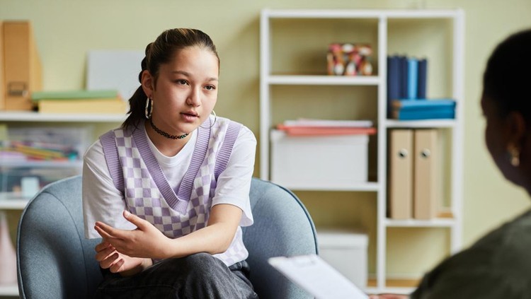1405791770 Teenage girl sitting on armchair and talking about her problems to psychologist during consultation at office