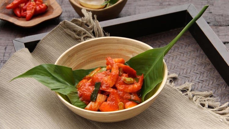 643525172 A popular dish of stir-fried shrimps in red chili paste from Padang, West Sumatra. Plated in a bamboo bowl lined with a broad green leaf. Arranged on a wooden tray that has been lined with casually-laid place mat. A small wooden saucer of red chili peppers and a wooden bowl of spoons are placed next to the tray. The dish is styled on a dark color, rustic wooden table.
