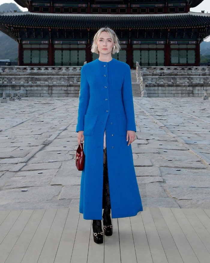 Saoirse Ronan bergaya sartorial dalam balutan mantel biru. Busana klasik tersebut ia beri statement lewat platform heels, stocking, dan tas Gucci Jackie warna merah. Foto: Instagram Gucci