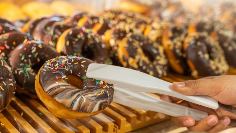 1165986686 Woman hand using clamp picking yummy chocolate donut at pastry shop