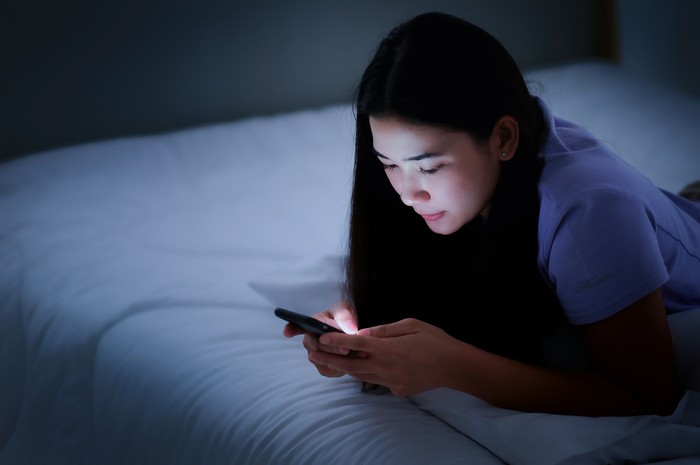 Asian young woman lying on the white bed and playing smartphones during night time. She is chatting with her friend. Using phone in low light is impact to eyes. Health and social Concept