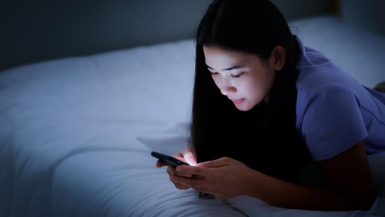 Asian young woman lying on the white bed and playing smartphones during night time. She is chatting with her friend. Using phone in low light is impact to eyes. Health and social Concept