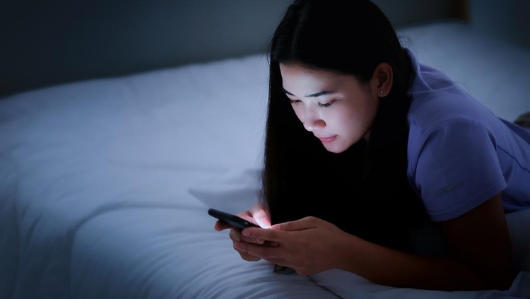 1215086216 Asian young woman lying on the white bed and playing smartphones during night time. She is chatting with her friend. Using phone in low light is impact to eyes. Health and social Concept