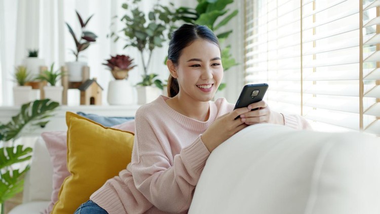 Young attractive beautiful casual asia female lady student look at cellphone play social media sit at sofa couch living room feel relax comfort carefree with green houseplant at cozy home apartment.