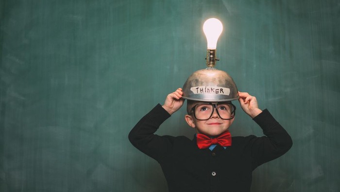 A young nerd boy dressed in retro 1980s attire, with bow tie and eyeglasses, wears a light bulb idea invention machine to help him think of the next big idea.