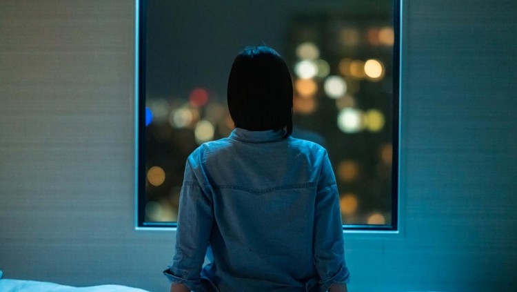 1388406488 A rear view of a woman sitting alone on a bed in room and looking through the window at night.