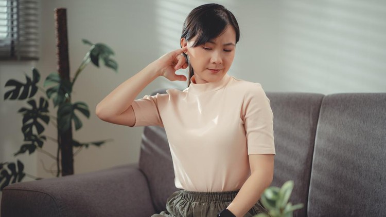 ilustrasi telinga tersumbat Asian woman itching from ear inflammation and putting a finger into her ear sitting on sofa in living room at home.