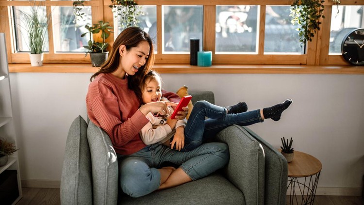 Cute little girl and her mother sitting in armchair, surfing the net together, using smartphone.