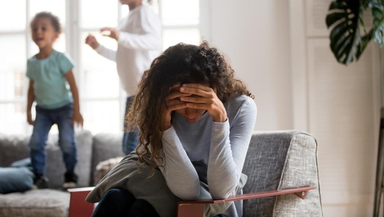 1051381408 Black single frustrated woman hold her head with hands sitting on chair in living room, playful kids jumping on couch on a background. Tiredness, depression difficult to educate children alone concept