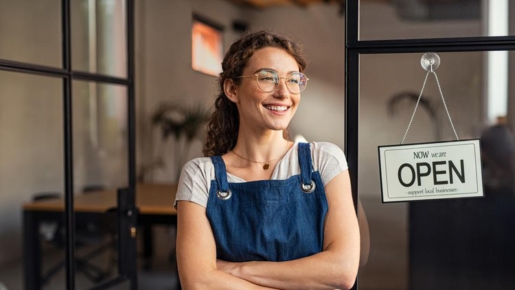 Portrait of happy waitress standing at restaurant entrance while looking outdoor. Portrait of young business woman attend new customers in her coffee shop. Smiling small business owner showing open sign in her shop while leaning against the door.