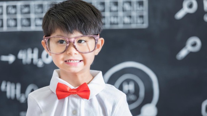 Cute Asian schoolboy has a smile on his face while standing in front of a chalkboard in his science classroom. He is wearing large glasses and a red bow tie.