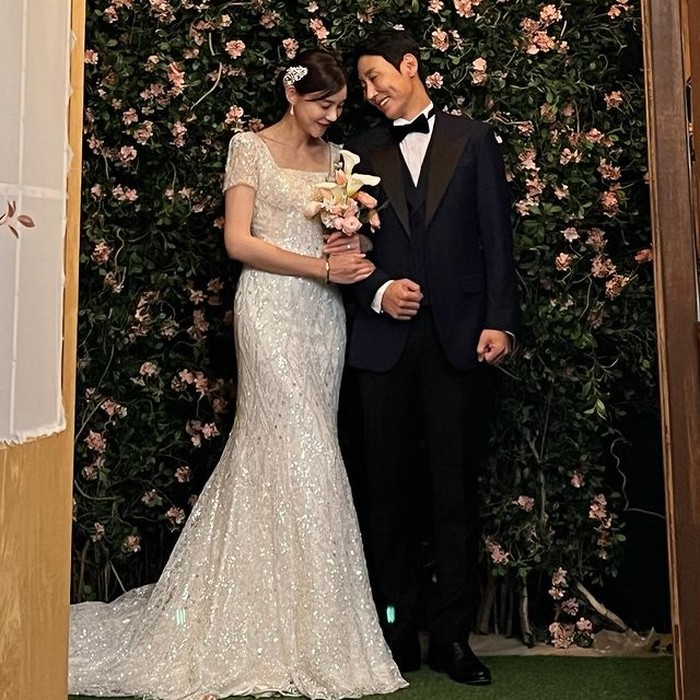 Dilatarbelakangi ornamen bunga-bunga yang menawan, Kim Na Young (Cha Ye Ryun) menggandeng erat Lee Tae Ho (Kim Young Hoon) dalam drama 'Battle for Happiness' sambil menggenggam buket bunga berwarna merah muda./ Foto: instagram.com/chayeryun