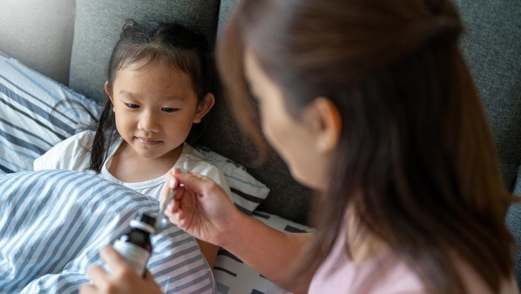 An Asian mom pouring cough syrup medicine into clear spoon to her daughter at bedroom,