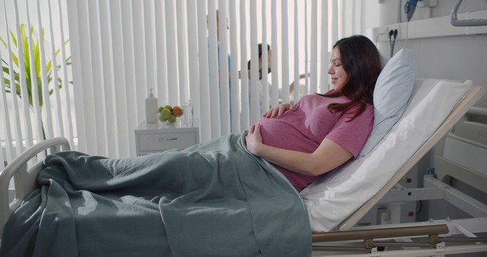 Pregnant young woman lying on the bed in delivery room. Side view of happy pregnant female resting on bed in hospital ward. Healthcare and pregnancy concept