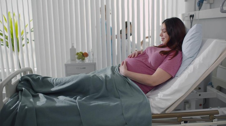 Pregnant young woman lying on the bed in delivery room. Side view of happy pregnant female resting on bed in hospital ward. Healthcare and pregnancy concept