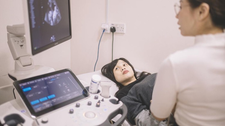 A pregnant female of Asian decent, lays out on an exam table as a technician conducts her ultrasound.  She is dressed casually and has her belly exposed as she looks to the screen to see her baby.