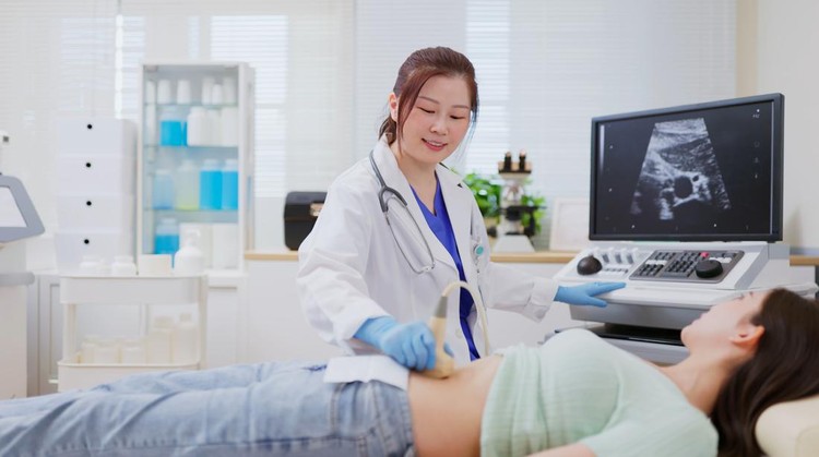 A pregnant female of Asian decent, lays out on an exam table as a technician conducts her ultrasound.  She is dressed casually and has her belly exposed as she looks to the screen to see her baby.