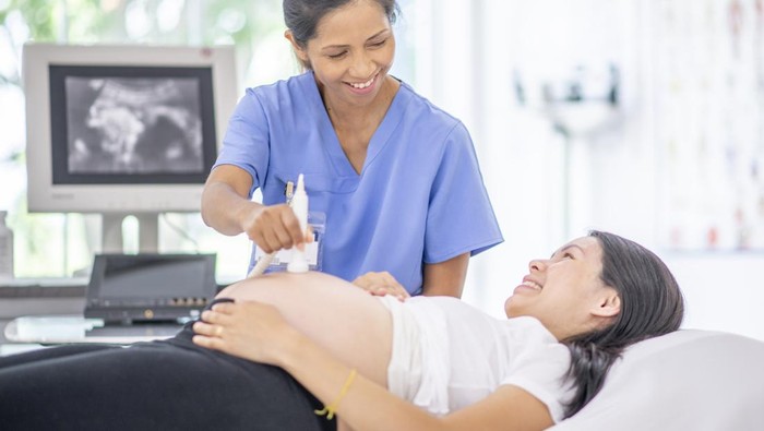 A pregnant female of Asian decent, lays out on an exam table as a technician conducts her ultrasound.  She is dressed casually and has her belly exposed as she looks to the screen to see her baby.
