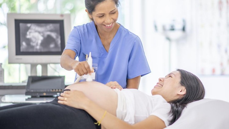 A pregnant female of Asian decent, lays out on an exam table as a technician conducts her ultrasound.  She is dressed casually and has her belly exposed as she looks to the screen to see her baby.
